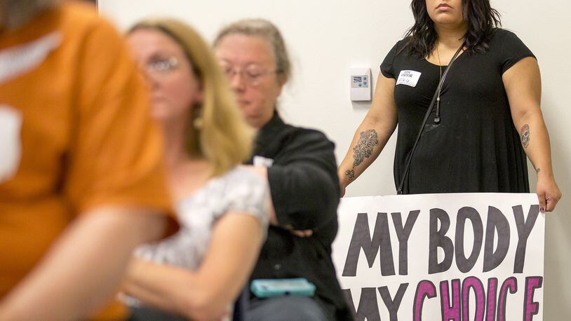 Lexi Cardenas stands in the back of the room holding a sign during an August hearing about proposed rules that aborted and miscarried fetuses not used for scientific research must be buried or cremated.