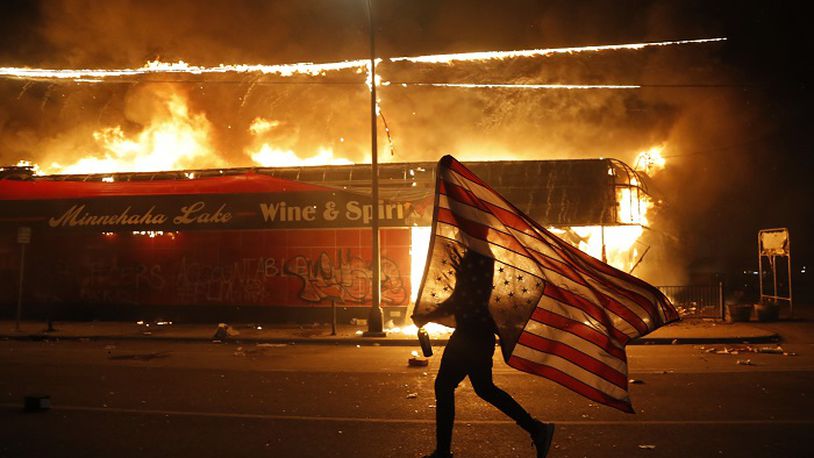 A protester carries a U.S. flag upside, a sign of distress, next to a burning building Thursday, May 28, 2020, in Minneapolis. Protests over the death of George Floyd, a black man who died in police custody Monday, broke out in Minneapolis for a third straight night. (AP Photo/Julio Cortez)