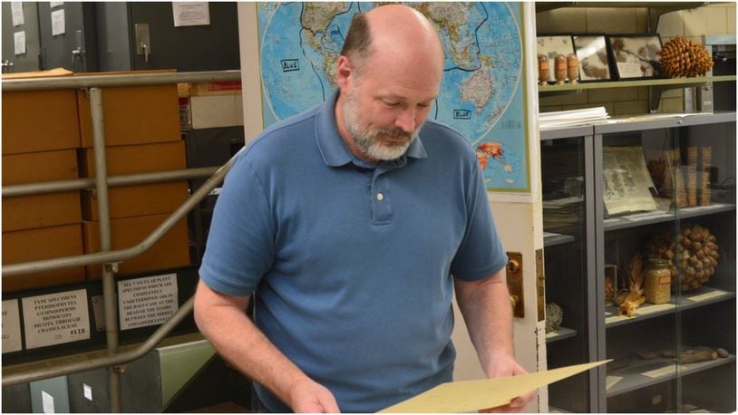 Dr. Michael Vincent looks at a plant specimen mounted on paper which is being prepared for sending to a researcher. Behind him is the map of the world showing the color-coding scheme used for labeling the samples stored in the three floors of cabinets accessed by the stairs through the door. CONTRIBUTED/BOB RATTERMAN