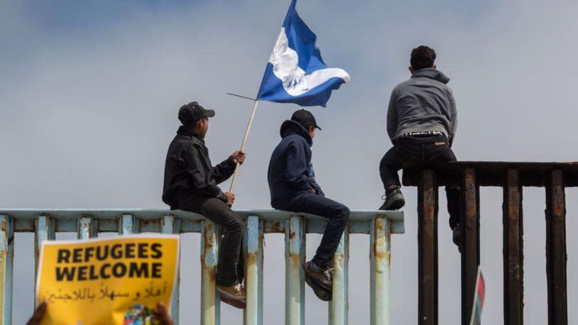 People hold a Honduran flag and look into the United States from atop a section of border fence as members of a caravan of Central American asylum seekers arrive in Tijuana, Mexico.