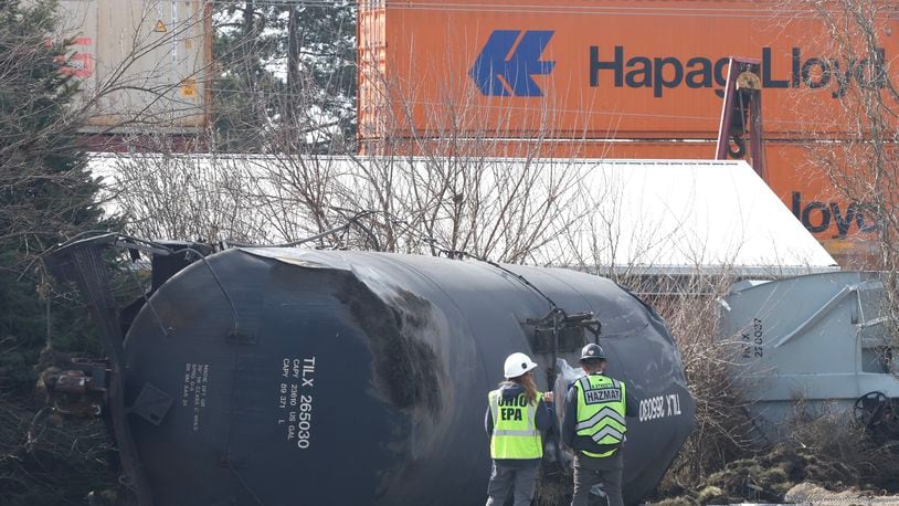 A HAZMAT team member and an Ohio EPA representatives stand by an overturned tank car as they watch another train travel down the tracks in the background Monday, March 6, 2023, in Springfield. A Norfolk Southern train derailed in Clark County near the fairgrounds on Saturday. BILL LACKEY/STAFF