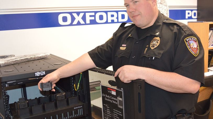 Police Chief John Jones holds one of the department s body worn cameras on top of the docking station which charges the units and also serves as a computer link for downloading the video. CONTRIBUTED/BOB RATTERMAN