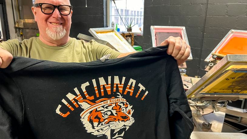Dondi Carder holds up one of their in-house designed and printed tiger themed Cincinnati shirts at Unsung Salvage Design Company on Main Street in Hamilton. Unsung Salvage has had a hard time keeping up with demand for the shirts during the Bengals' playoff run.  NICK GRAHAM / STAFF
