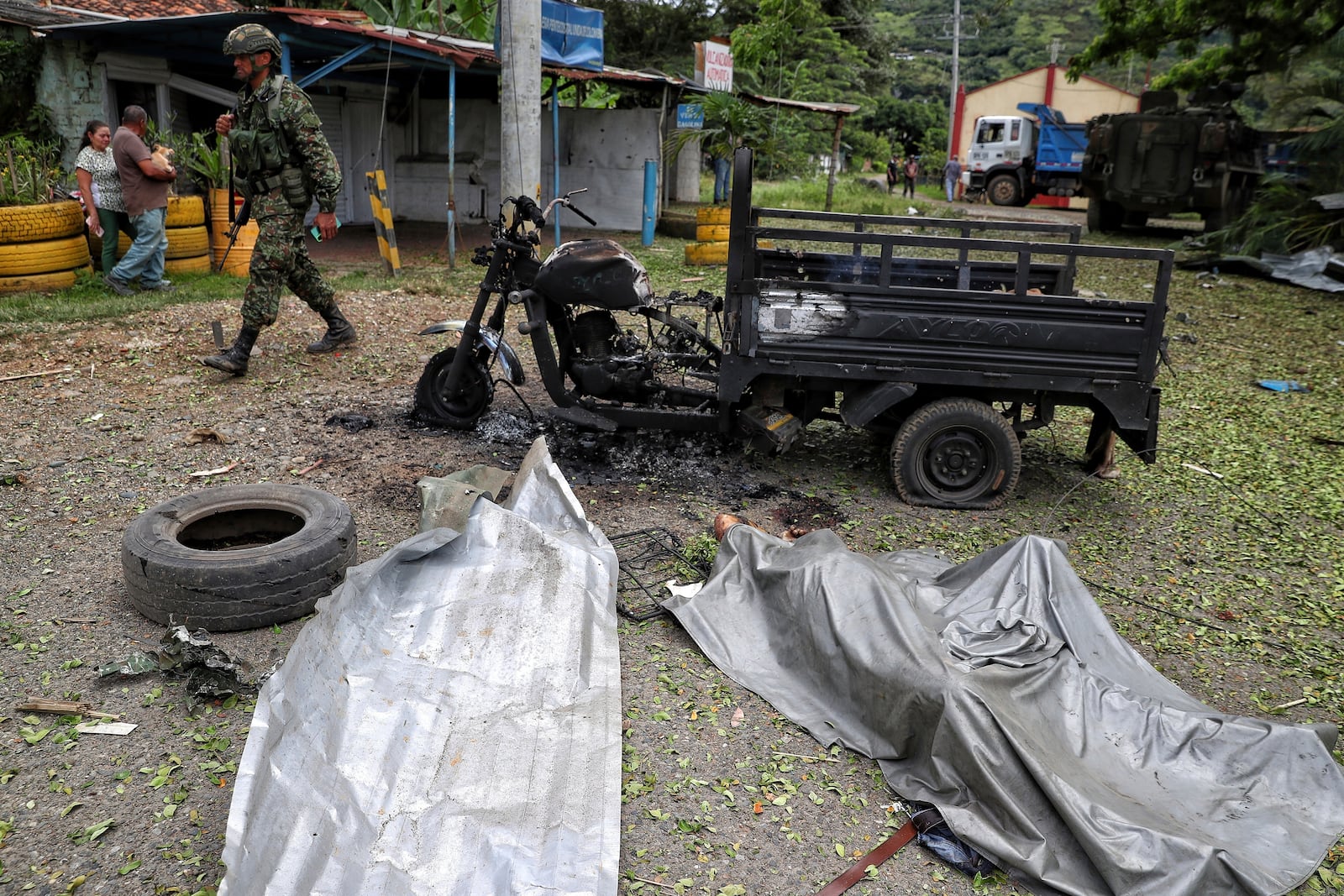 A soldier walks past the body of a man covered in plastic, right, and his charred motorcycle, after the person was killed by a bomb that exploded in Guachinte, Cauca Valley, Colombia, Tuesday, June 10, 2025. (AP Photo/Santiago Saldarriaga)