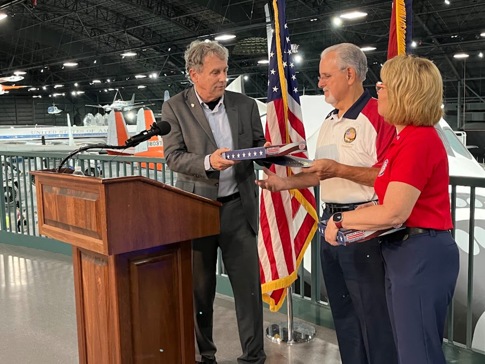 U.S. Sen. Sherrod Brown, D-Ohio, hands Tom Ritchie, president of the Dayton-Miami Valley Regional Labor Council, a flag that has flown over the U.S. Capitol on Wednesday, Aug. 7, 2024 at the National Museum of the U.S. Air Force. Next to Ritchie is Kim Frisco, executive director of the Montgomery County Veterans Services Commission. THOMAS GNAU/STAFF