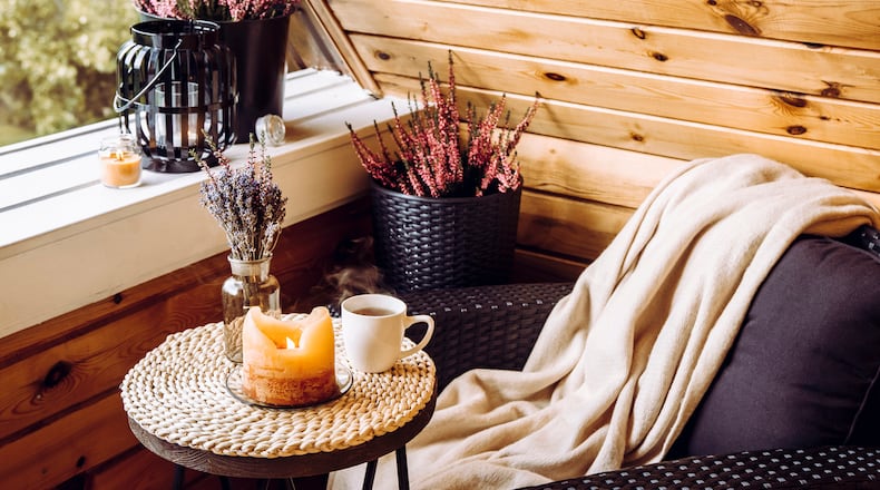 A tiny wooden cabin balcony is seen with heather flowers in a pot, lavender in a bottle vase, a candlelight flame and a soft beige plaid waiting on comfortable garden furniture chair. iSTOCK/COX