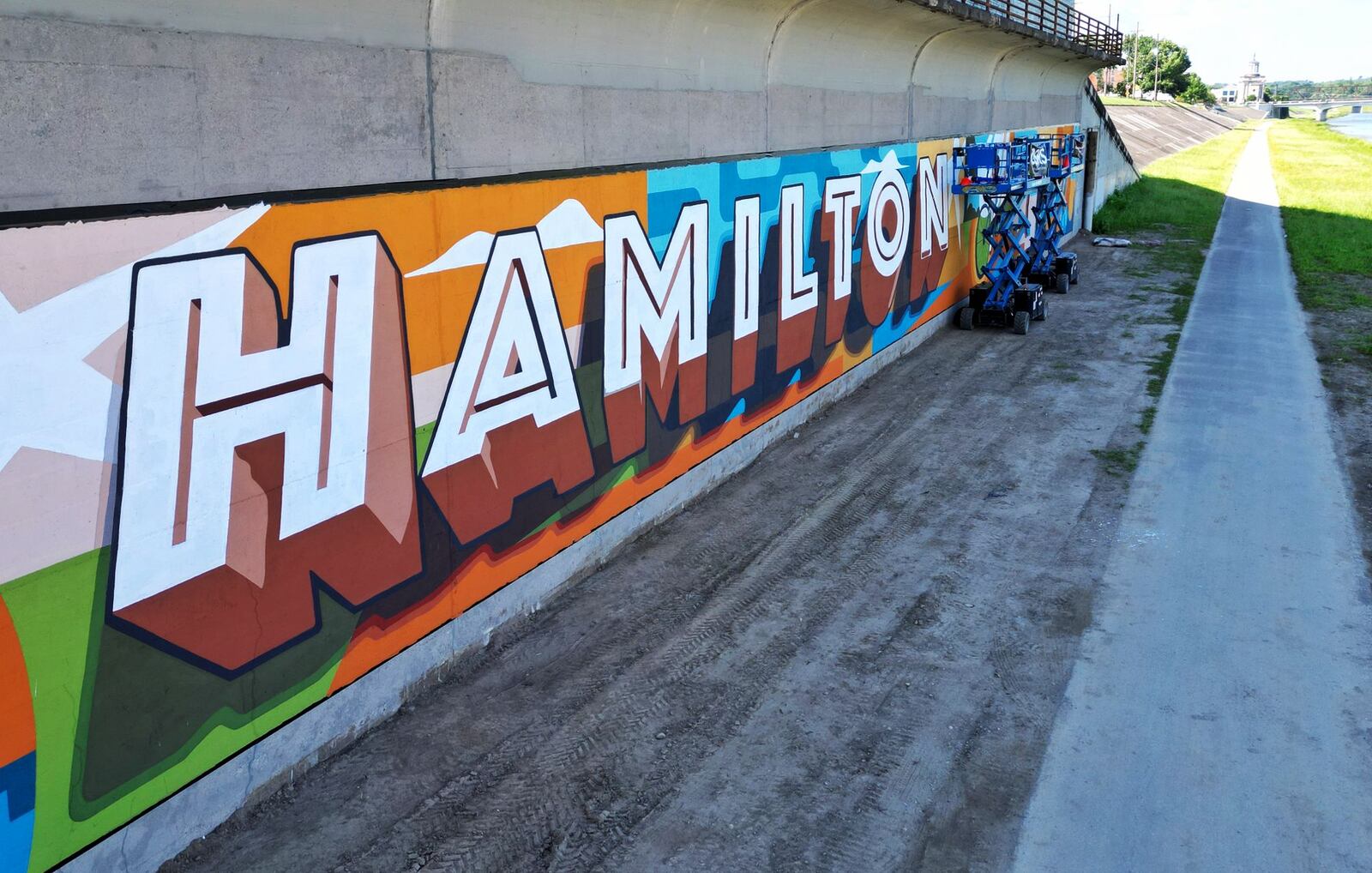 Work is nearing completion on the second-largest StreetSpark mural since the program started nearly a decade ago. The winning design, titled Tourism Series: Hamilton, is by the Cincinnati and Louisville-based mural company and art collective Often Seen Rarely Spoken. The mural is aling the bike path by the Great Miami River across from Spooky Nook Sports Champion Mill in Hamilton. NICK GRAHAM/STAFF