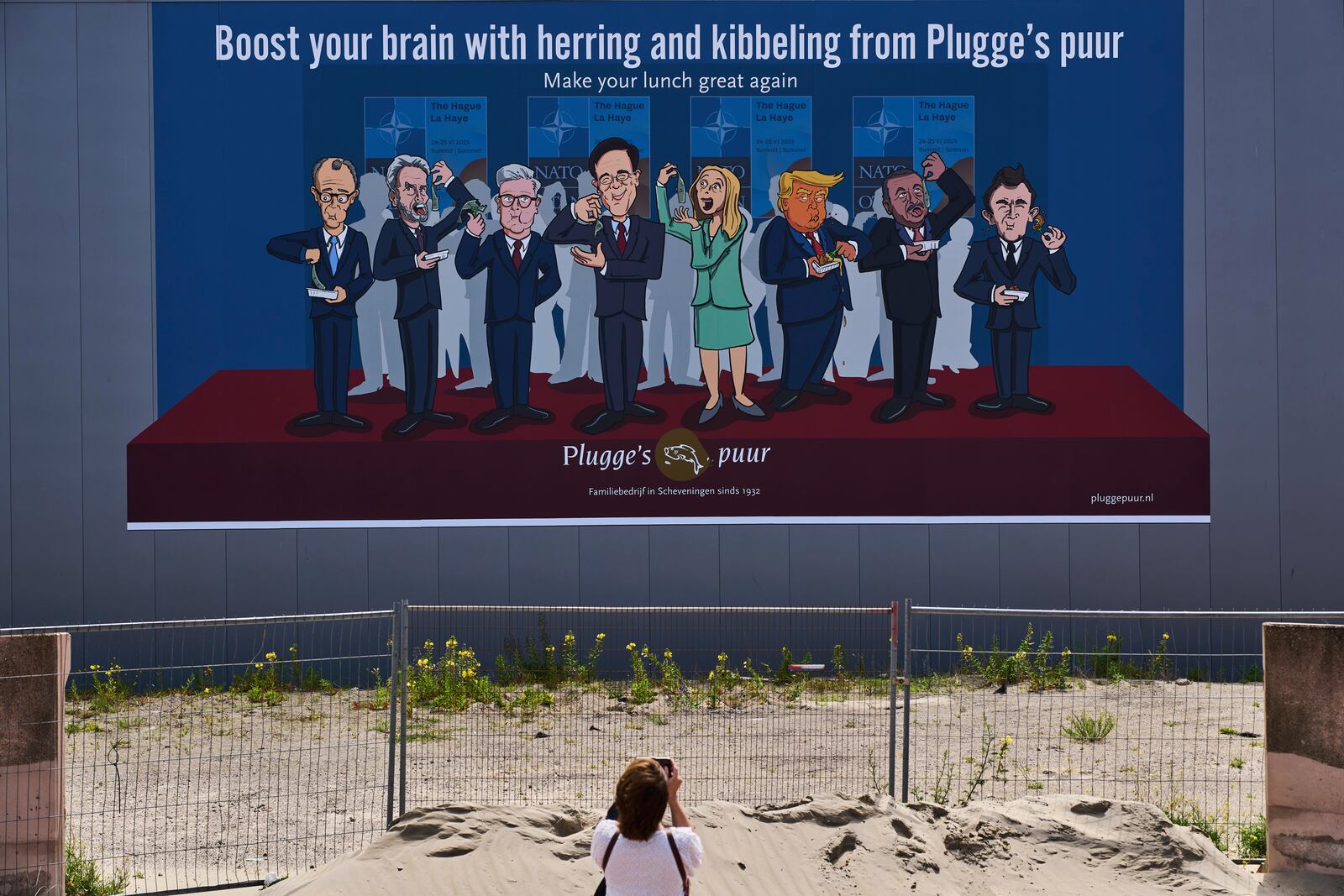 A woman takes a photo of a drawing showing World leaders from left, German Chancellor Friedrich Merz, Dutch Prime Minister Dick Schoof, British Prime Minister Keir Starmer, NATO Secretary General Mark Rutte, Italy's Prime Minister Giorgia Meloni, U.S. President Donald Trump, Turkish President Recep Tayyip Erdogan and French President Emmanuel Macron, eating herring and fried fish in The Hague, Netherlands, Sunday, June 15, 2025. (AP Photo/Peter Dejong)
