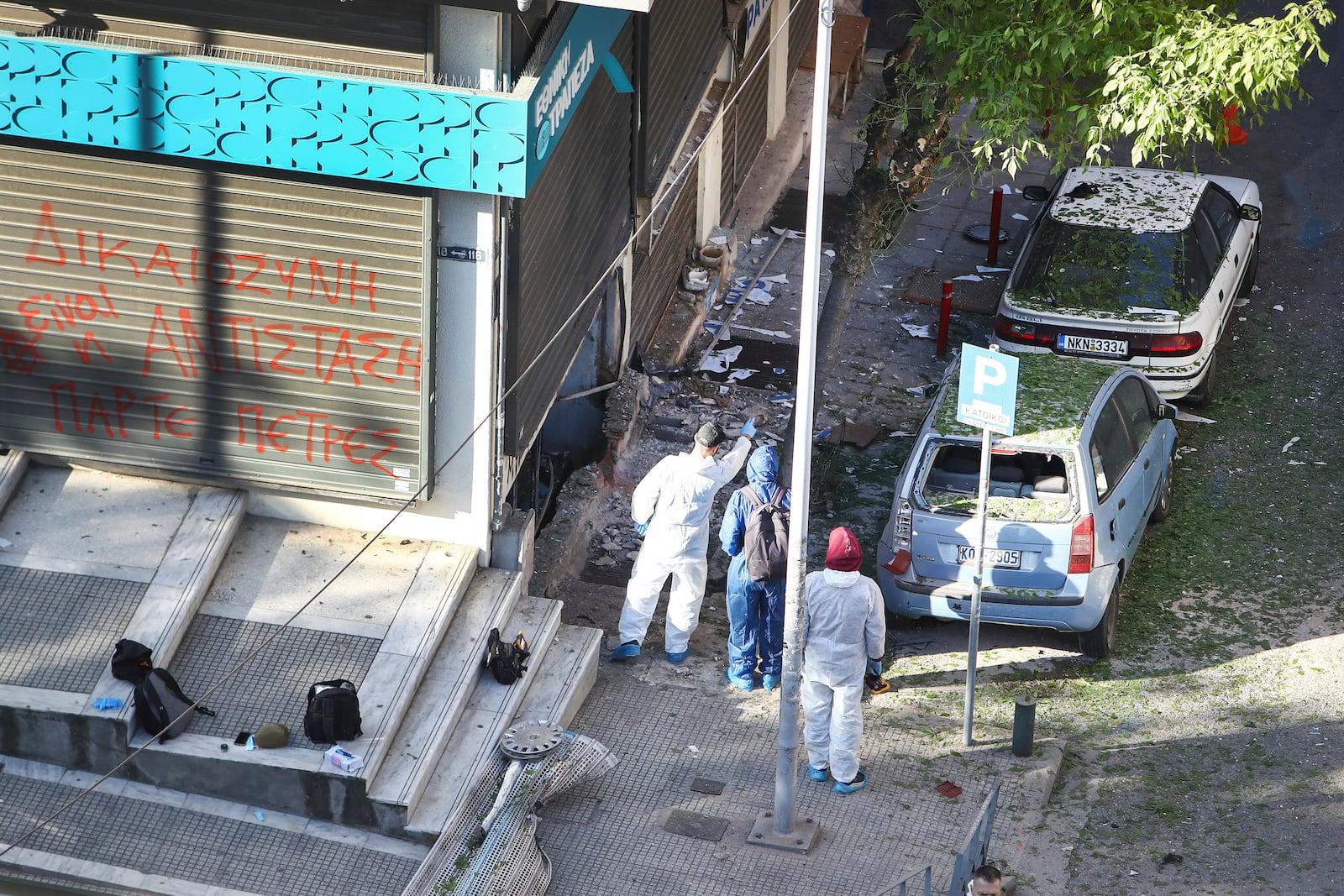 Greek police forensic experts search the area where a 38-year-old woman was killed early Saturday when a bomb she was carrying exploded in her hands outside a bank branch, local police said, in Thessaloniki, northern Greece, May 3, 2025. On the fence of the bank branch reads : "Justice is to resist. Take stones". (AP Photo/Giannis Papanikos)