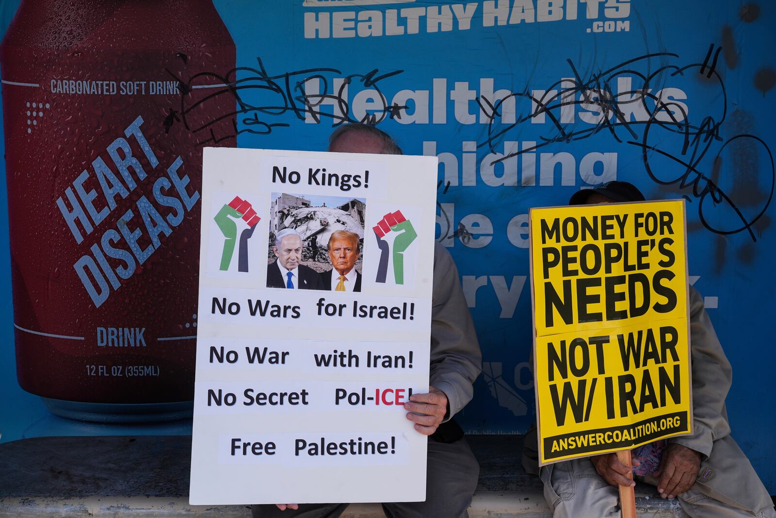 Two anti-war demonstrators hold signs at a bus stop outside the Wilshire Federal Building in Los Angeles Sunday, June 22, 2025, following U.S. airstrikes on Iran's nuclear facilities. (AP Photo/Jae C. Hong)