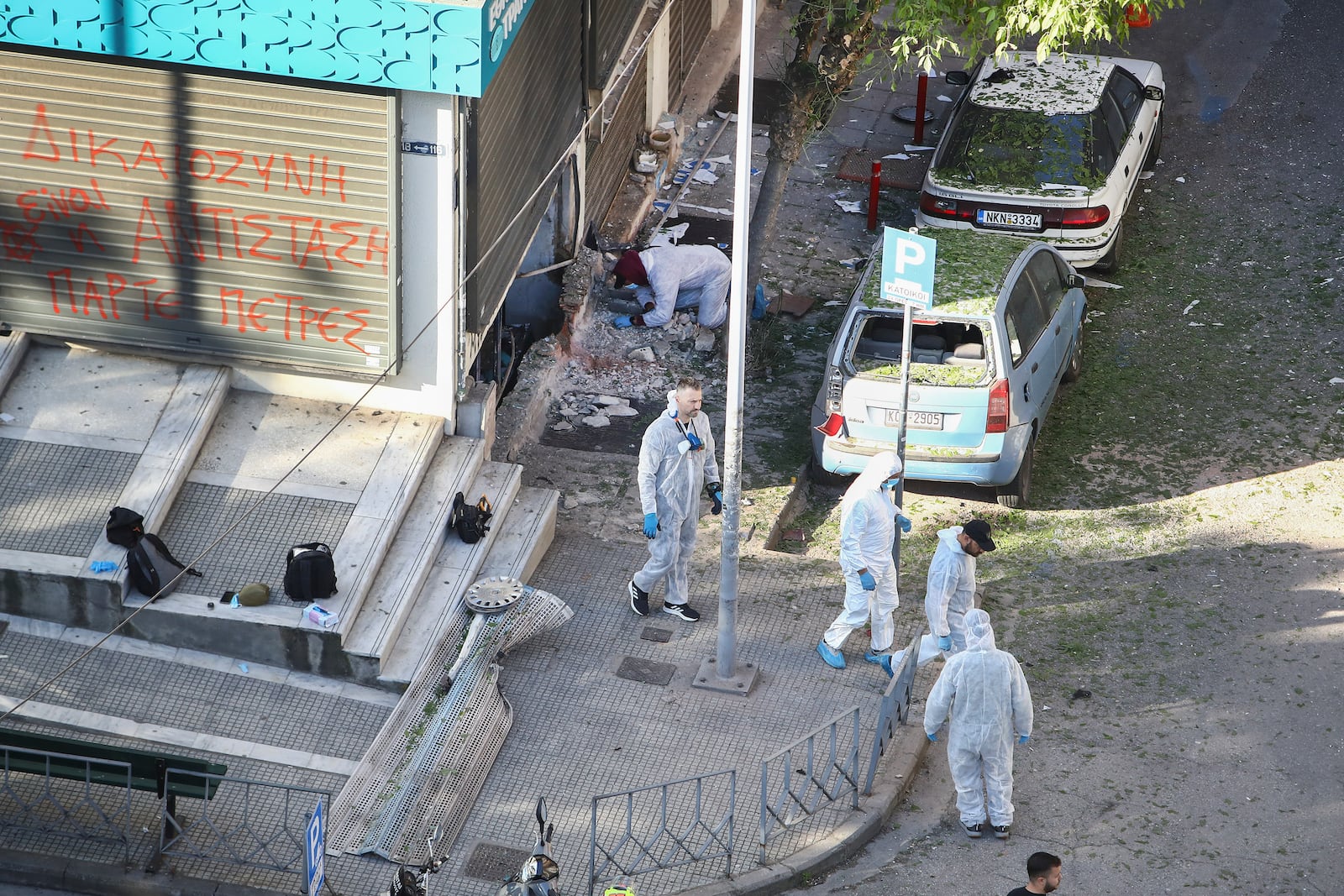Greek police forensic experts search the area where a 38-year-old woman was killed early Saturday when a bomb she was carrying exploded in her hands outside a bank branch, local police said, in Thessaloniki, northern Greece, Saturday, May 3, 2025. On the fence of the bank branch reads : "Justice is to resist. Take stones". (AP Photo/Giannis Papanikos)