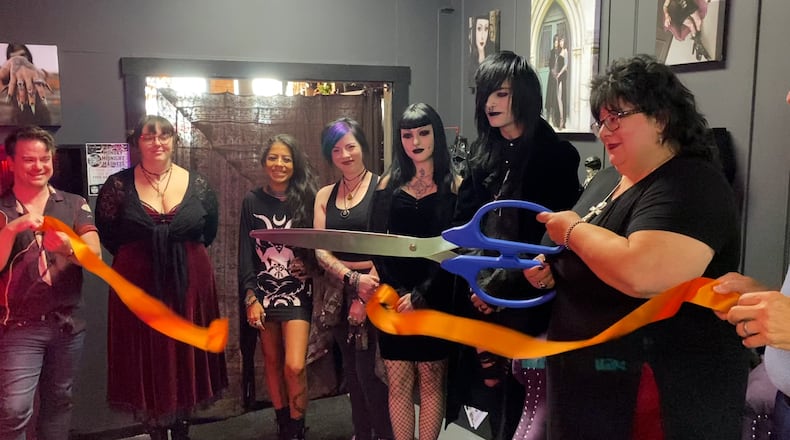 Dark Side Of The Moon is bringing something different to Main Street. The Trad Goth boutique features clothing items and accessories in a goth style. It's located in the back of Seraph By The River, 118 Main St., Hamilton, and both stores are owned by Dawn Singleton. Pictured, with Singleton holding the oversized scissors, is the ribbon cutting of the new boutique on Friday, July 19, 2024. PROVIDED/TVHamilton