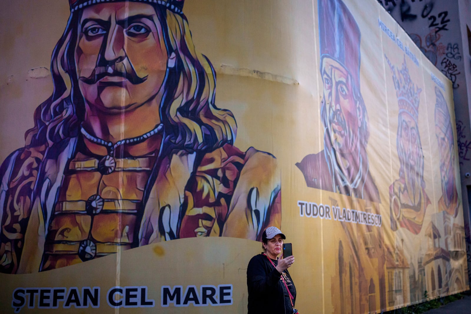 A woman holds up a mobile phone backdropped by depictions of Romanian historical figures outside the headquarters of the Alliance for the Unity of Romanians (AUR) shortly before polls close in the first round of the country's presidential election redo in Bucharest, Romania, Sunday, May 4, 2025. (AP Photo/Andreea Alexandru)