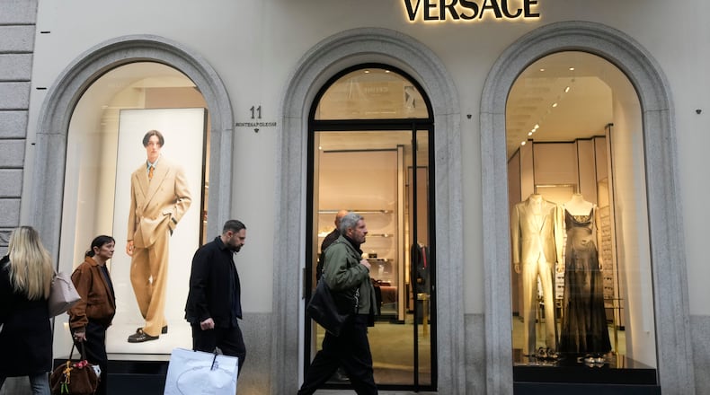 A man walks with a Prada shopping bag in front of a Versace shop, at the Montenapoleone luxury fashion street, in Milan, Italy, Tuesday, Dec. 2, 2025. (AP Photo/Luca Bruno)