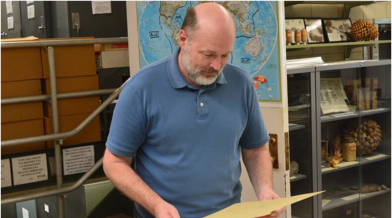 Dr. Michael Vincent looks at a plant specimen mounted on paper which is being prepared for sending to a researcher. Behind him is the map of the world showing the color-coding scheme used for labeling the samples stored in the three floors of cabinets accessed by the stairs through the door. CONTRIBUTED/BOB RATTERMAN