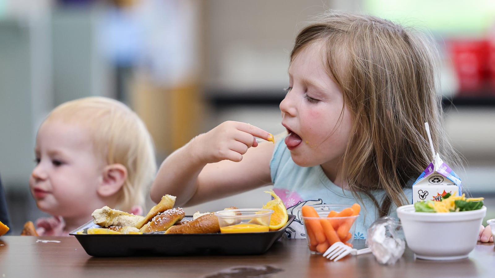 Emmy (right) licks cheese sauce off a finger during a summer lunch program organized by Kettering City Schools at Greenmont Elementary School. She was eating at the final lunch of the summer, which was on Friday, Aug. 1. Their mother Dana took her along with younger sister Violet (left) and older sister Ava (unpictured) to the program once or twice a week. She said it was a good socialization opportunity for the kids, who often played at the school's playground after eating. They were also joined on Friday by Dana's sister and the children's aunt, Abby. BRYANT BILLING / STAFF