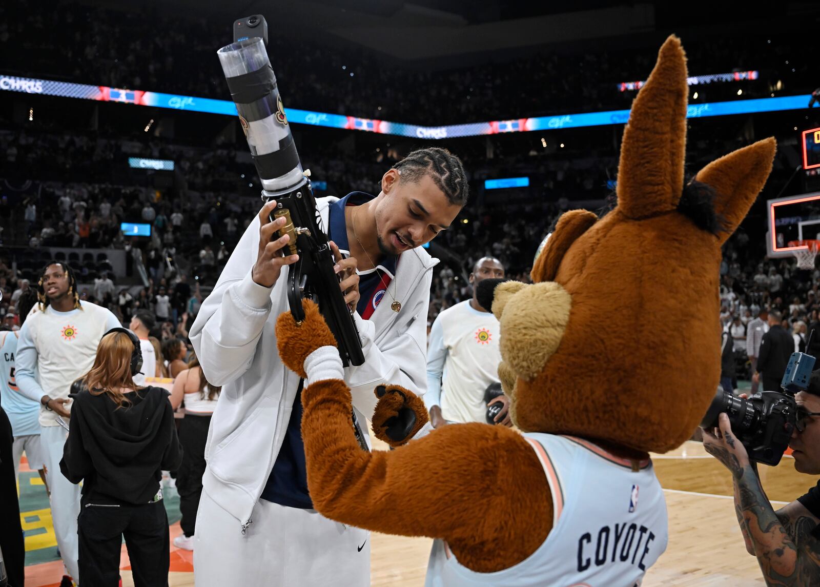 The San Antonio Spurs Coyote, right, teaches Spurs forward Victor Wembanyama how to use the tee shirt cannon after an NBA basketball game against the Toronto Raptors, Sunday, April 13, 2025, in San Antonio. (AP Photo/Darren Abate)