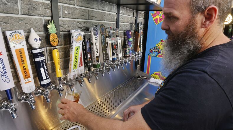 Pinball Garage owner Brad Baker pours a Municipal Brew Works beer, crafted by a fellow Hamilton business, Thursday, July 21, 2022, in Hamilton. Many businesses in Hamilton support each other in various ways. NICK GRAHAM/STAFF