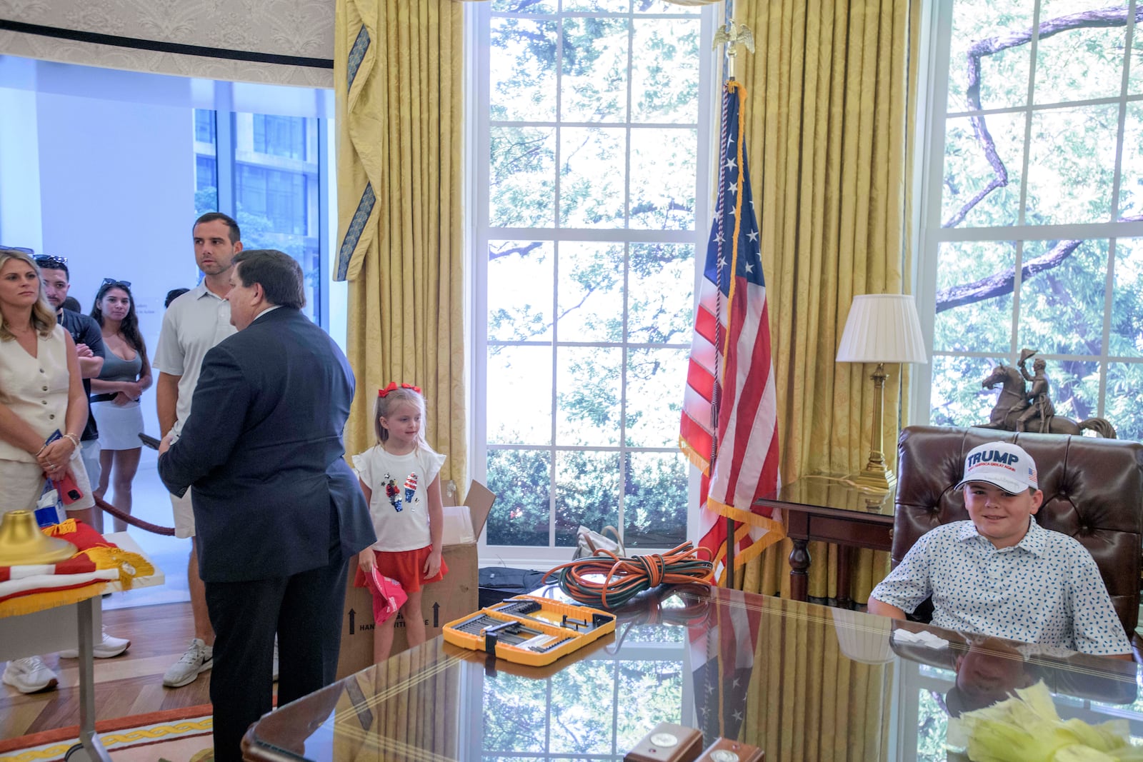 John Michael Cannette,10, right, of Sumrall, Miss., sits behind the Resolute desk as White House Historical Association President Stewart McLaurin, left, talks to a group of tourists, during work to transition a replica of the White House Oval Office from the days of former President Joe Biden with President Donald Trump's decor, at the White House Historical Association in Washington, Wednesday, July 23, 2025. (AP Photo/Rod Lamkey, Jr.)