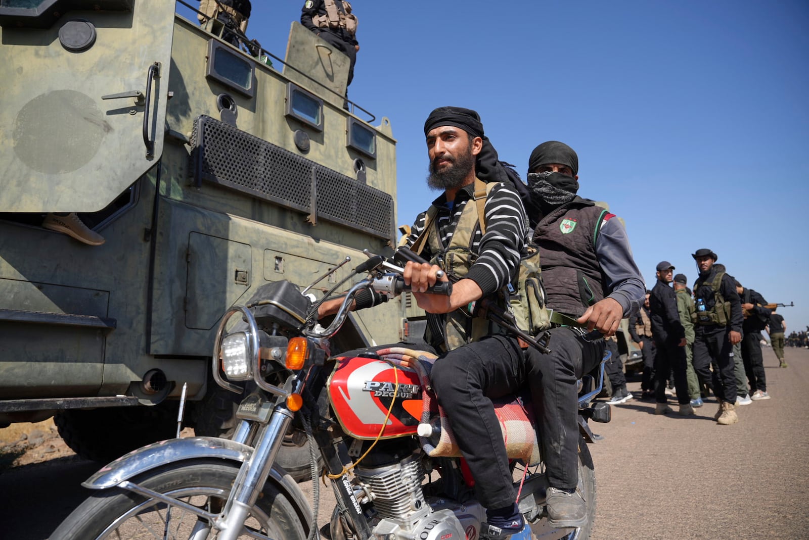 Two fighters from the Sunni Bedouin clans, ride their motorcycle as they pass by Syrian government security forces deployed on the outskirts of the Sweida province where clashes erupted between Druze militias and Sunni Bedouin clans, southern Syria, Monday, July 14, 2025. (AP Photo/Malek Khattab)