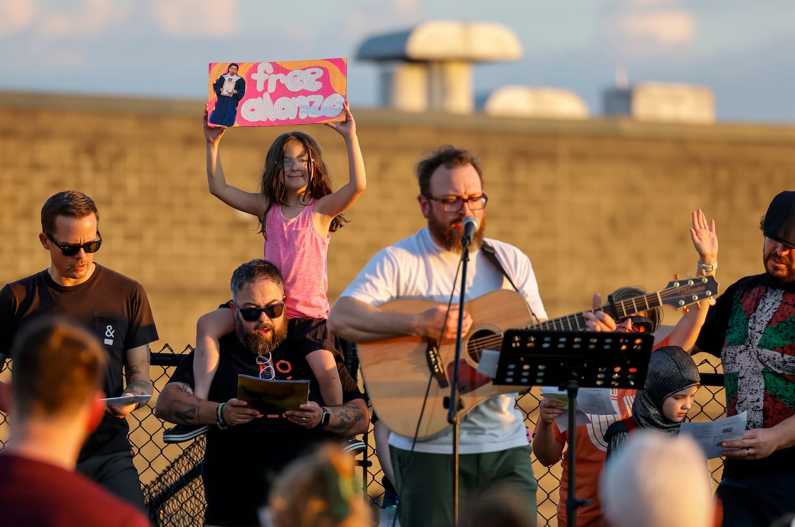 Maggie Heizer, 8, holds a sign on the shoulders of her father, Josh Heizer, as Tim Ferguson plays guitar during a candlelight vigil of over 100 faith leaders and advocates for Alonzo Tomas Mendez and other immigrants being held in Butler County Jail Tuesday, July 1, 2025 just outside the fence of the Butler County Sheriff's Office in Hamilton. NICK GRAHAM/STAFF