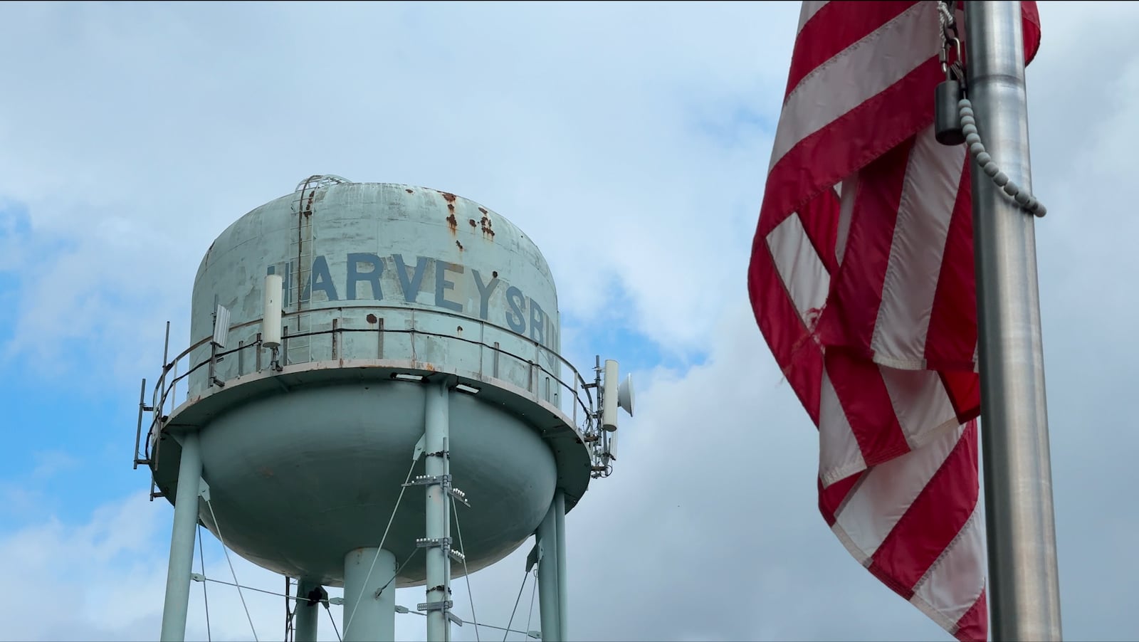 A weathered blue water tower with the word “Harveysburg” painted on it, photographed beside a U.S. flag on a flagpole.