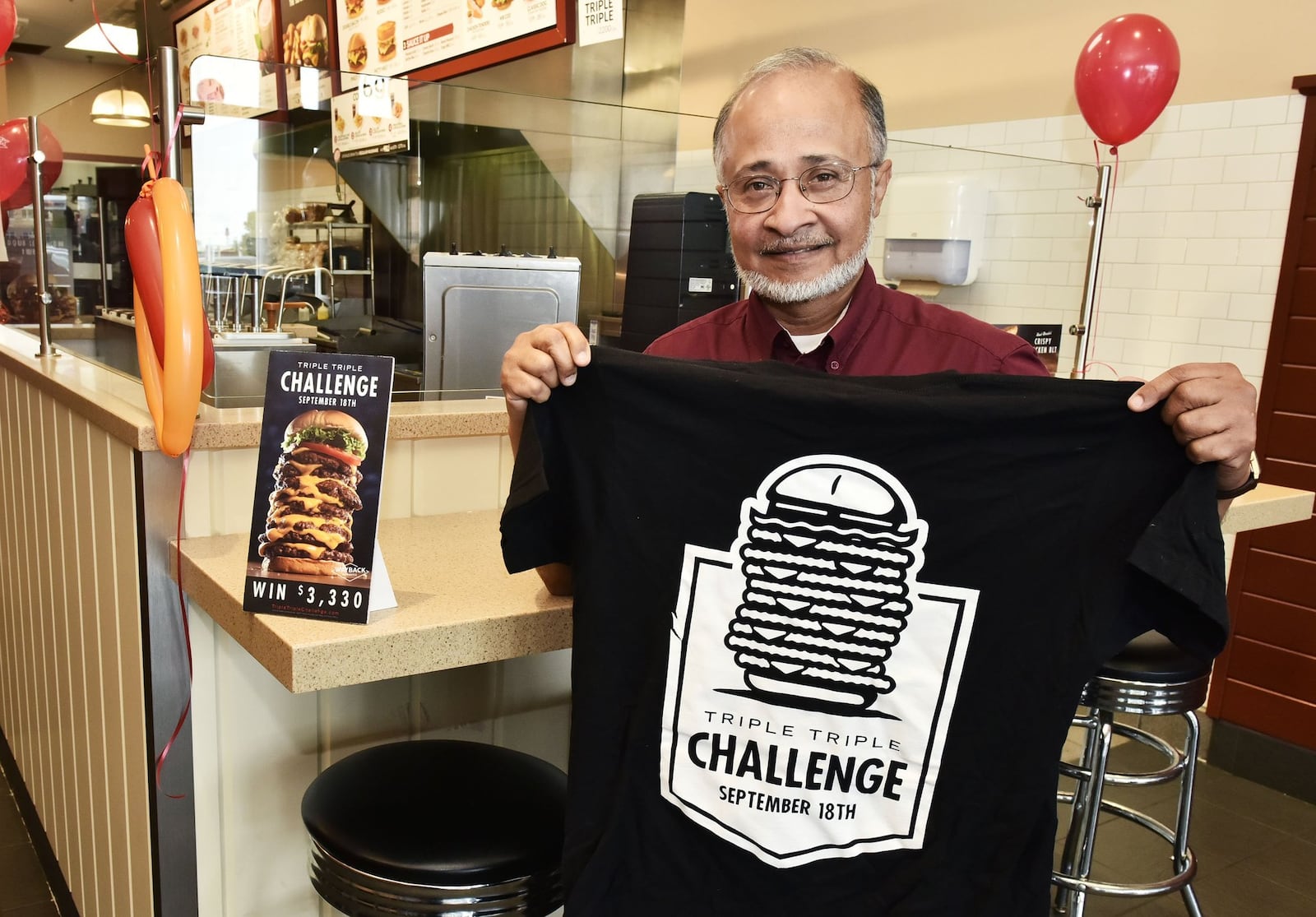 Nayeem Aziz, franchise owner of Wayback Burgers in West Chester Township, holds a shirt competitors of the franchise’s 10th annual Triple Triple Challenge will get if they try their luck at eating the gigantic nine-patty burger with nine slices of American cheese topped with lettuce, tomato and the bun. The competitive eating winner will take home $3,330 cash prize if they finish it in the the fastest time. The competition is a open nationwide at participating restaurants on National Cheeseburger Day. NICK GRAHAM/STAFF