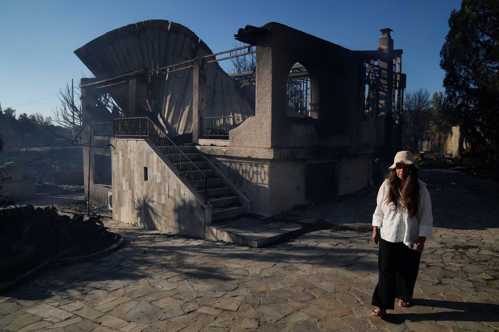 Lillian Tsertou walks in front of her burned down house in Thymari, south of Athens, Thursday, June 26, 2025, as authorities evacuate five locations during Greece's first summer heatwave with temperatures nearing 40 degrees Celsius (104 Fahrenheit). (AP Photo/Thanassis Stavrakis)