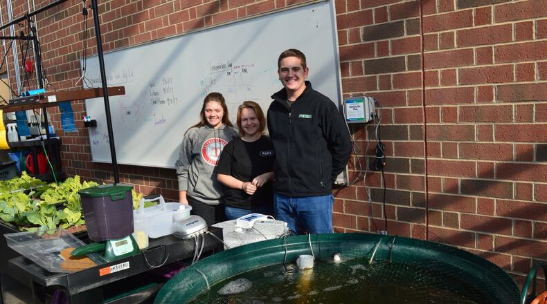 Talawanda sophomore greenhouse managers, from left are Hannah Lightfield, Eliza Jones and Justin Beckner stand behind their aquaponics system. The tank in the foreground holds the fish and the nutrients from their feces then move through the hoses and pipes to the stands on the other side which grow plants with roots in the water and no dirt. The water recycles back to provide nutrients to the fish. CONTRIBUTED/BOB RATTERMAN